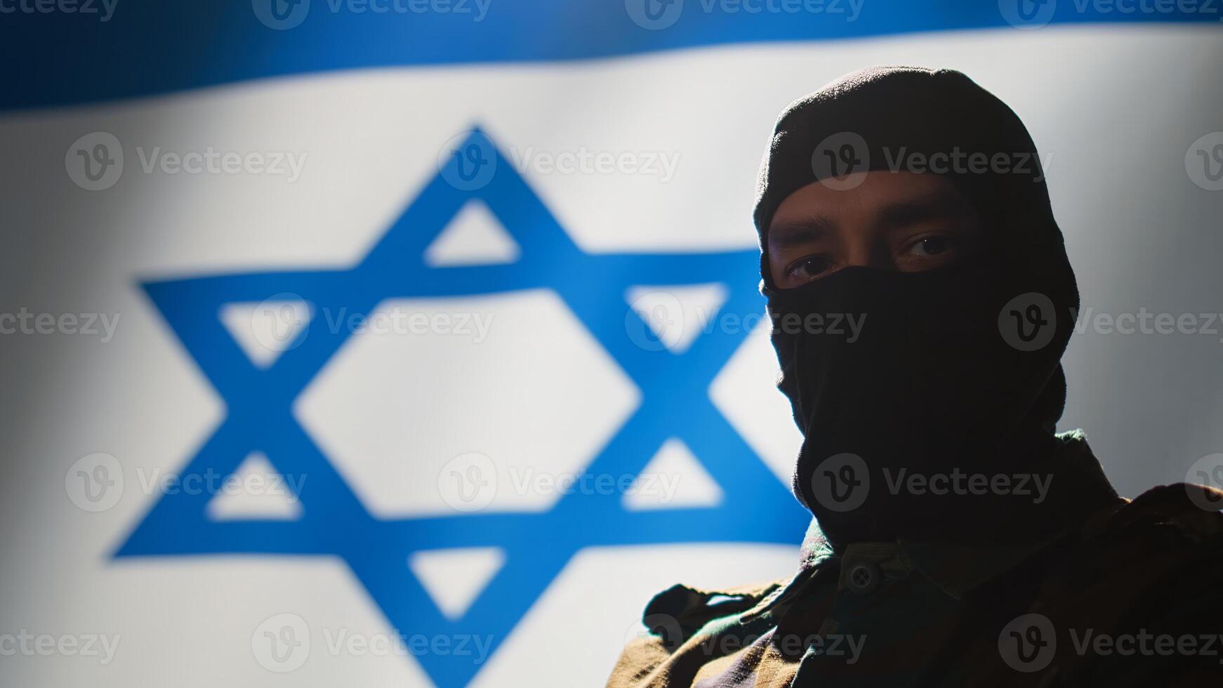 Mossad incognito spy wearing mask standing proud in front of Israel flag, feeling pride in serving country. Israeli secret agent in war control room crossing arms, representing country photo