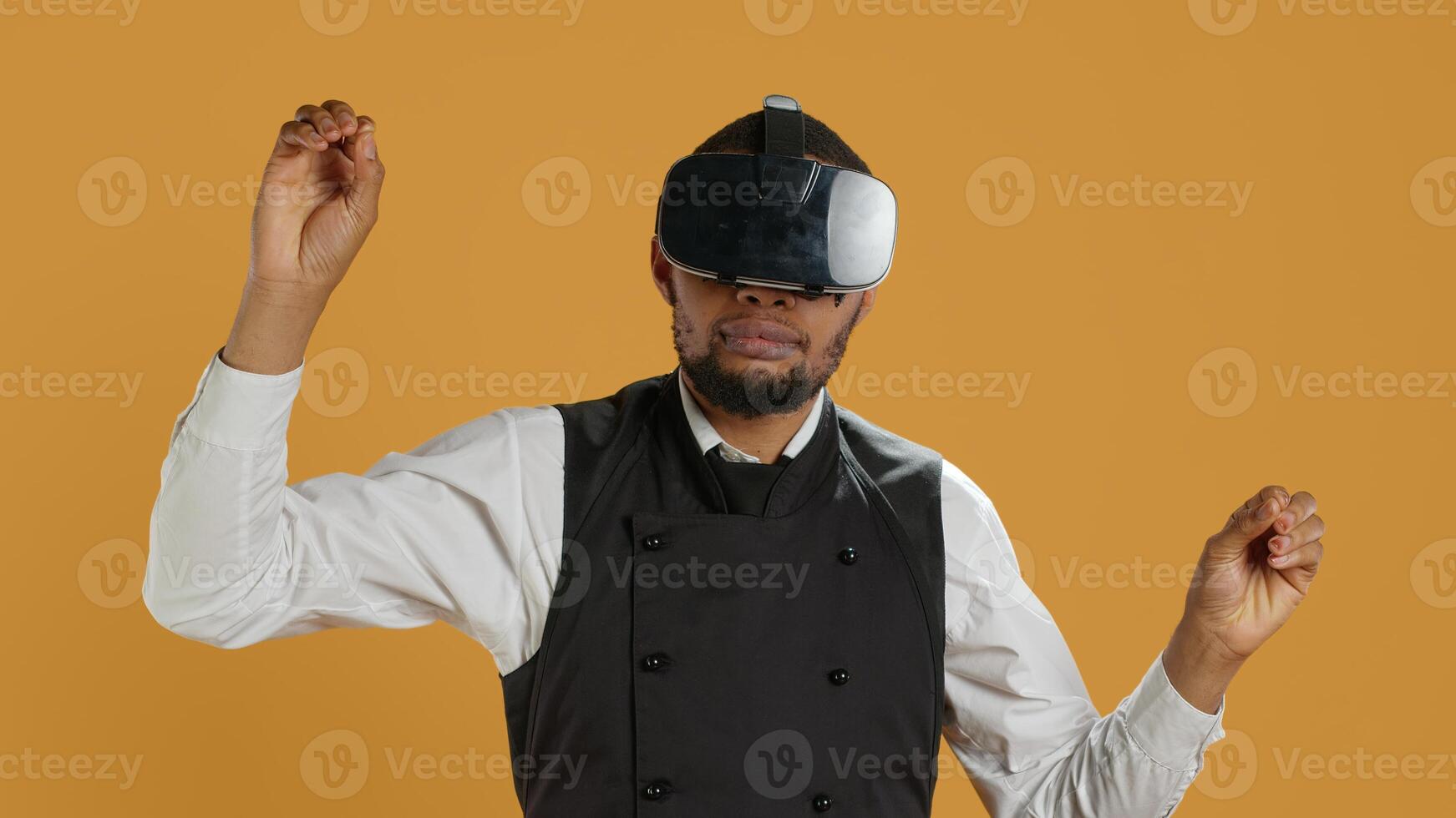 Restaurant waiter uses virtual reality headset with artificial intelligence, having fun with technology and vr glasses against yellow background. Employee with apron and uniform. Camera A. photo