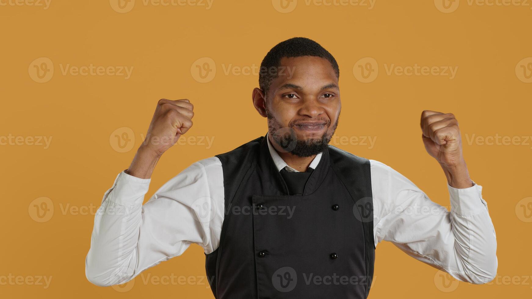 Waiter flexing his muscles and showing his strength in studio, acting tough while he wears his restaurant uniform and apron. Professional employee in service oriented industry. Camera A. photo