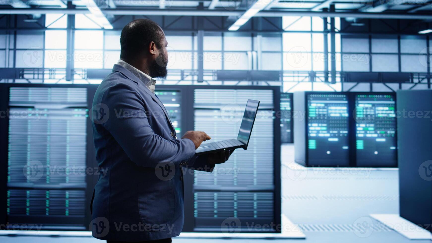 African american man walking in high tech facility, using laptop to patch network issues leading to slow data transfer rates. Data center worker fixing errors affecting supercomputers, camera B photo