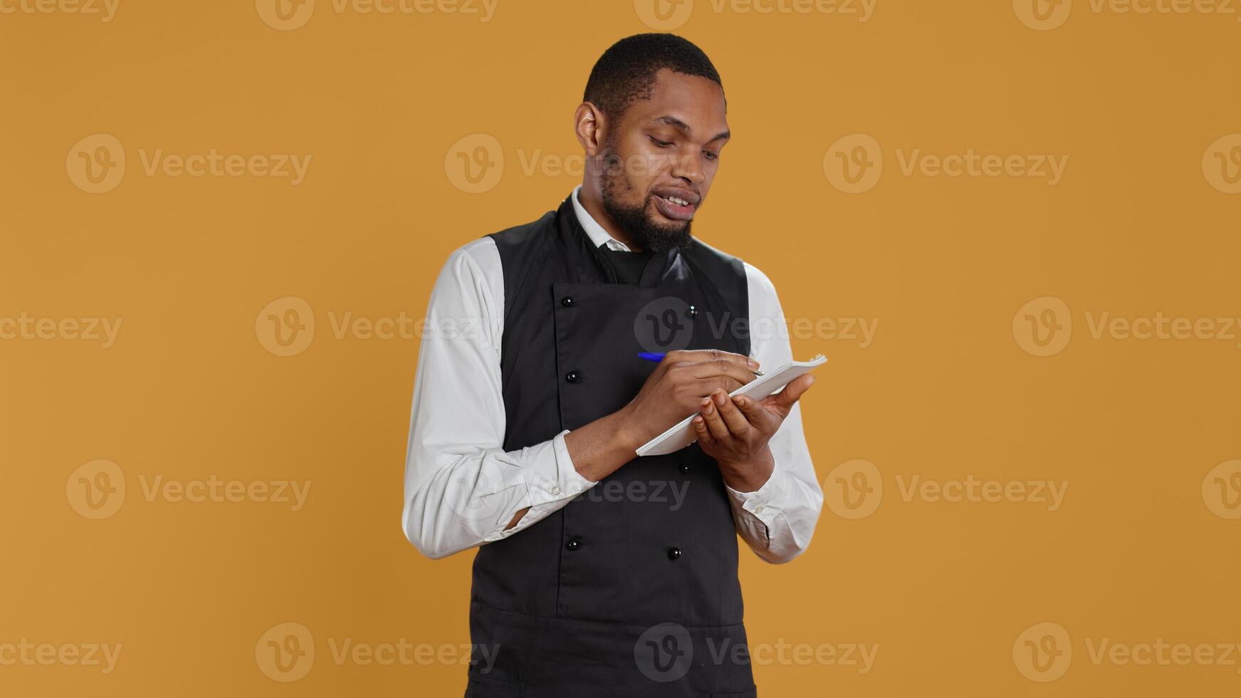 Skilled waiter with apron takes meal orders from clients on a notebook, writing down important information for customer satisfaction. Caterer butler working in service oriented industry. Camera B. photo