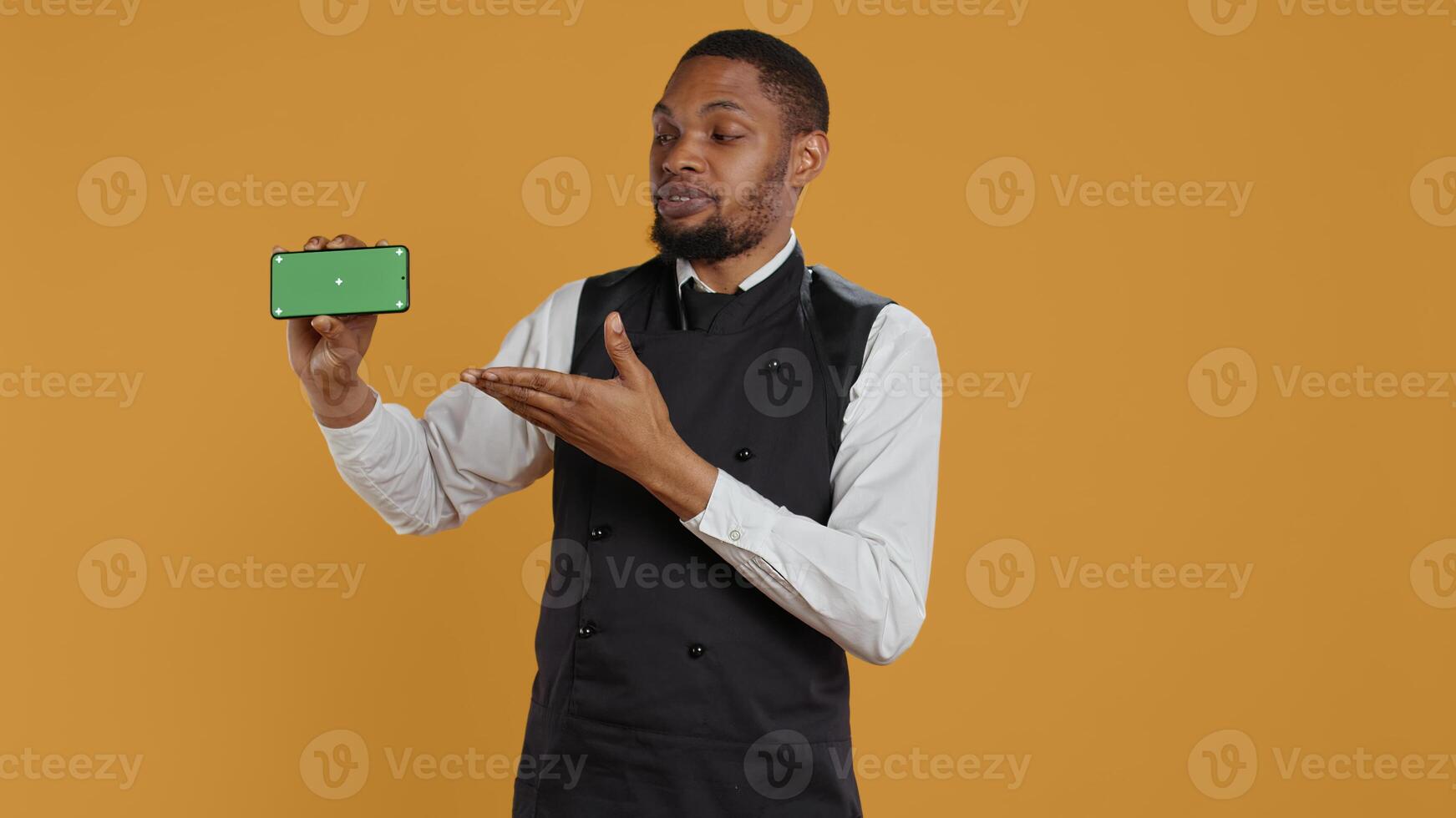 Waiter with professionalism holding a phone with isolated mockup display, working in dining and eatery industry. Friendly restaurant staff does customer oriented activity at his job. Camera B. photo