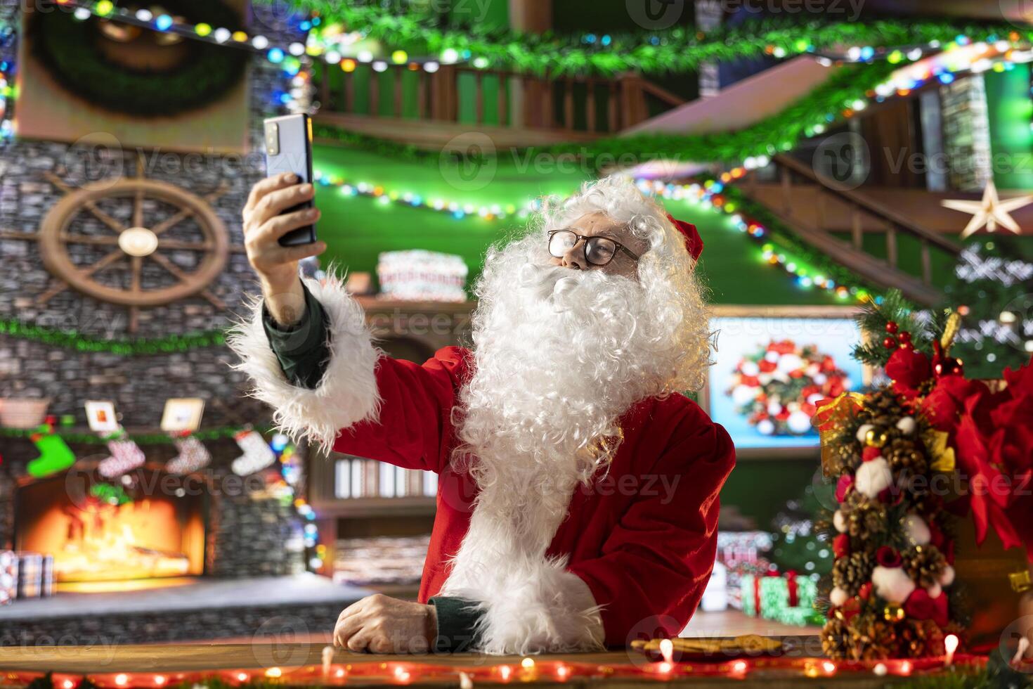 Old Father Christmas takes photos of himself in the festive red suit, having fun during the winter holidays to spread the x mas cheer. Santa taking pictures on his smartphone, modern tradition.