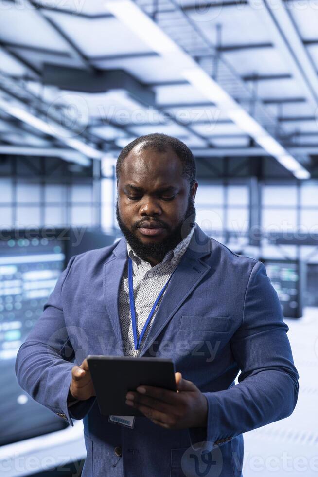 Technician walking in data center, using tablet to configure network infrastructure, such as routers, switches and firewalls. IT specialist in server hub looking for technical issues using device photo