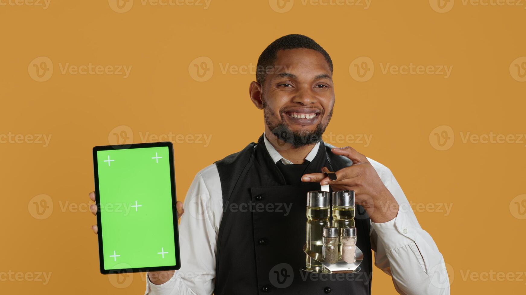 Male restaurant staff showing tablet with green screen against yellow background, carrying the cruet set with salt and pepper shakers used in serving clients. Service oriented job. Camera A. photo