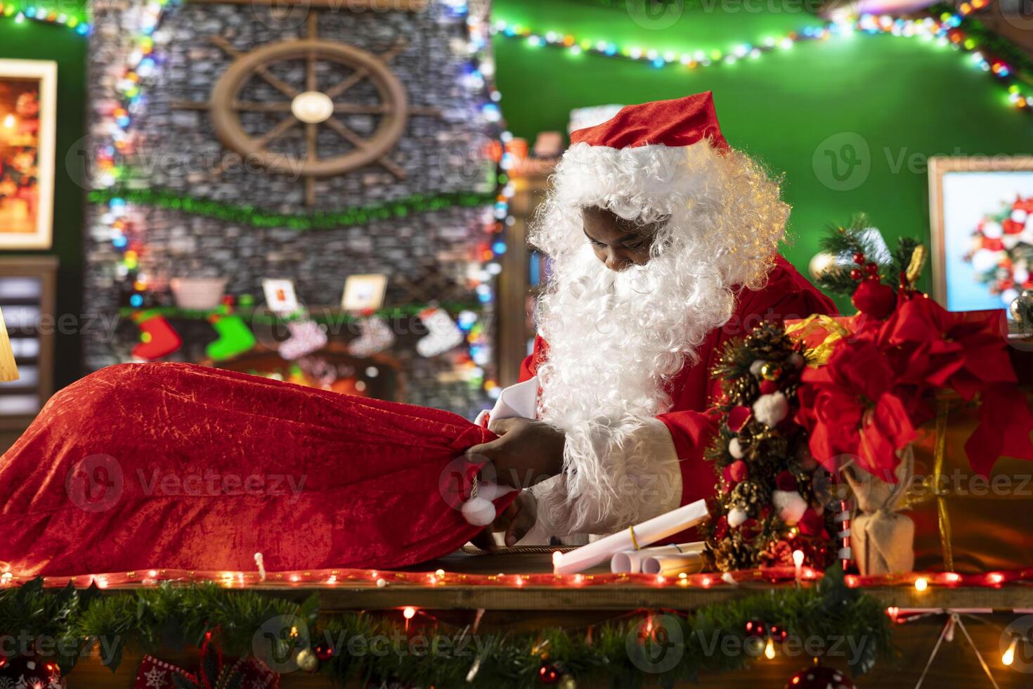 Black Santa filling his red sack with toys and presents for winter season holiday, spreading the Christmas spirit. Decorating gift boxes with wrapping paper, North Pole log cabin activity. photo