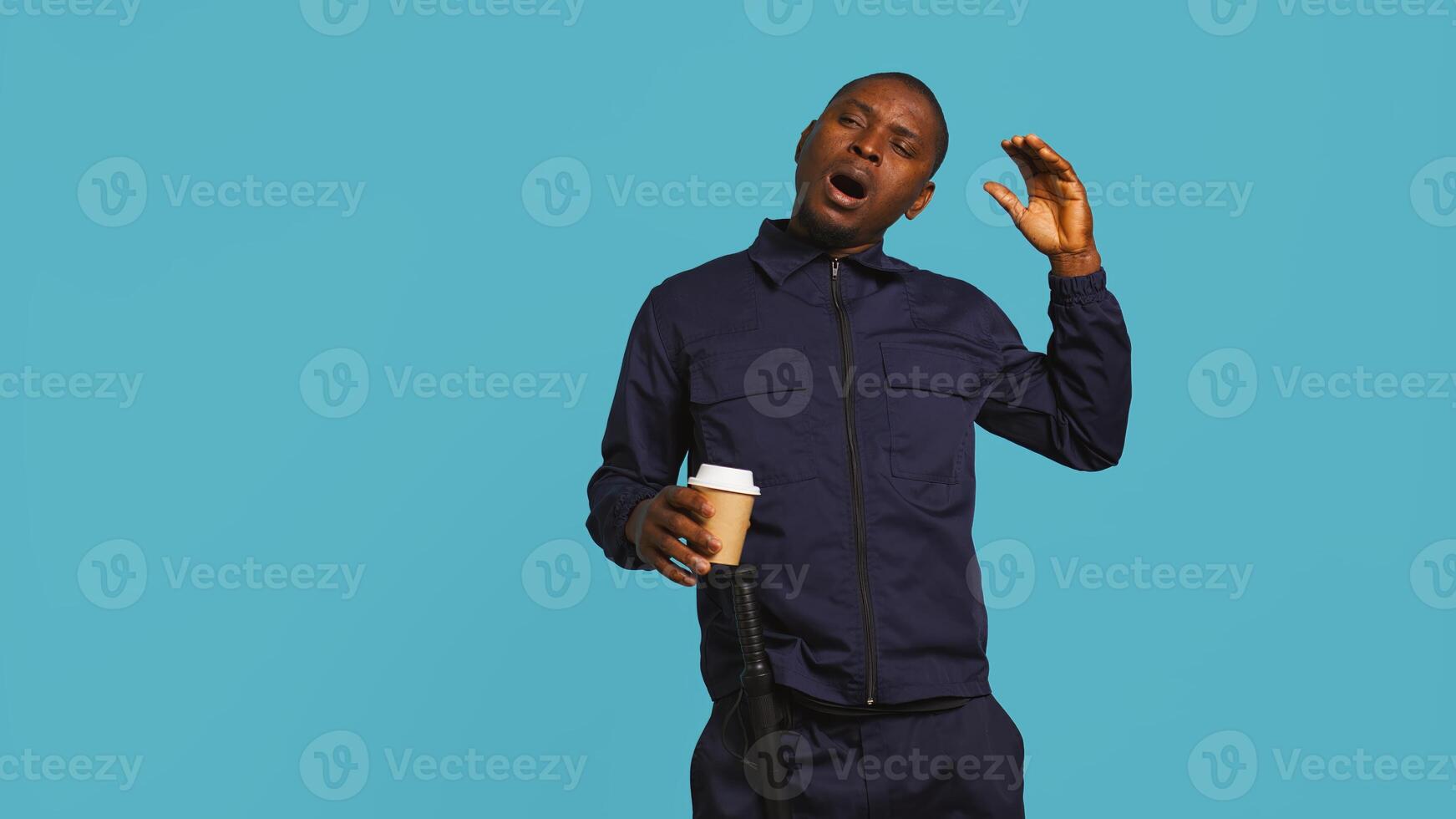 Portrait of fatigued security guard drinking coffee cup to remain energized while overseeing premises. Private law enforcement worker consuming caffeinated beverage, studio backdrop, camera B photo