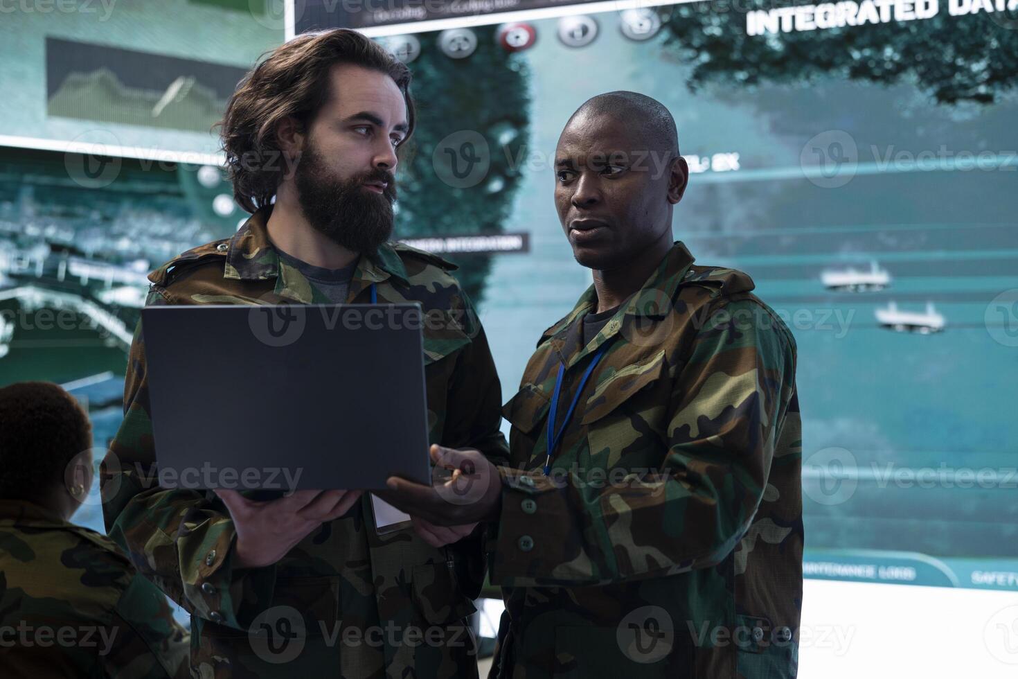Diverse team of IT operators comparing real time battlefield data from a laptop to a big screen, gather intelligence for future field missions. Collecting information from CCTV satellite surveillance. photo