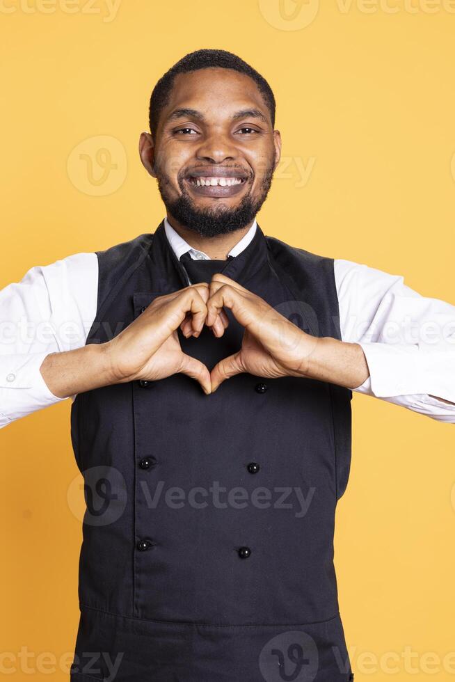 Waiter in apron showing a heart shape gesture against yellow background, expressing his sincere feelings and true love. Catering staff doing a romantic symbol, confident person. photo