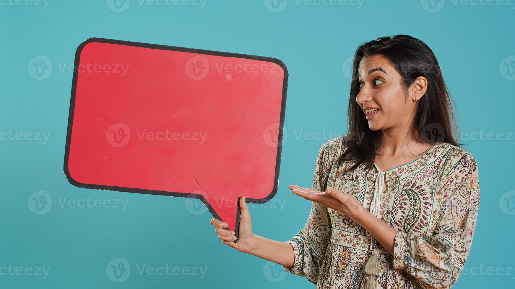 Smiling indian woman holding red speech bubble sign of empty copy space for message. Joyous person presenting thought bubble cardboard used as promotion concept, studio background, camera A photo
