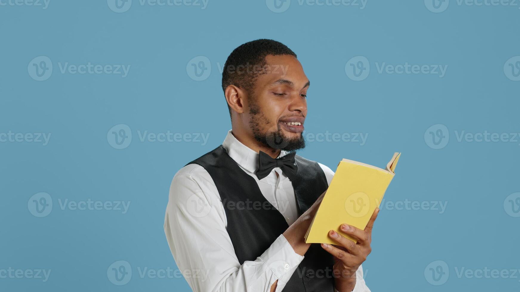 Young doorman hotelier reading a book on his work break in studio, hotel concierge in tuxedo against blue background. Bellhop resort personnel enjoys literature and lecture. Camera A. photo
