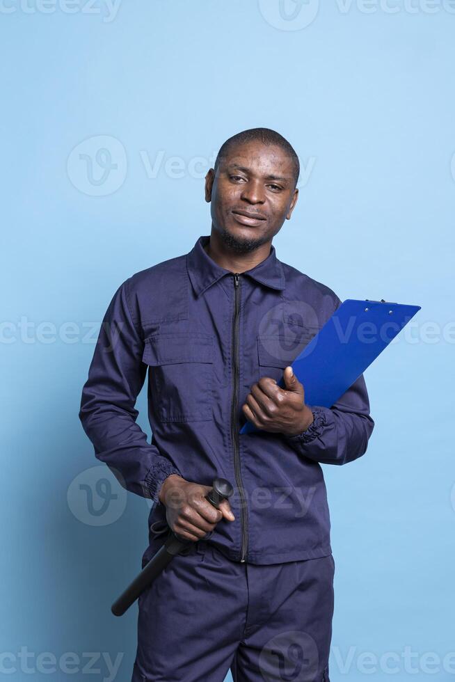 Portrait of confident bodyguard posing with his baton and papers, using a list of important activities to ensure safety and protection. Security agent stands against blue background. photo
