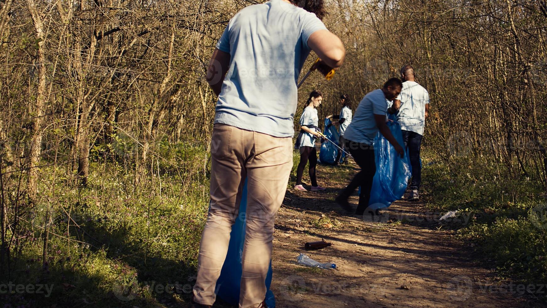 Team of volunteers cleaning the forest area from garbage and plastic, collecting rubbish with tongs and bags. Activists doing voluntary work to preserve natural habitat, illegal dumping. Camera B. photo