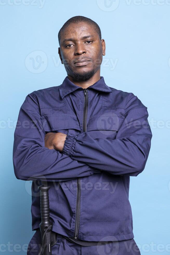 Portrait of african american guard posing with arms crossed against blue background, wearing his professional uniform with confidence and pride. Serious armed agent works on patrol service. photo