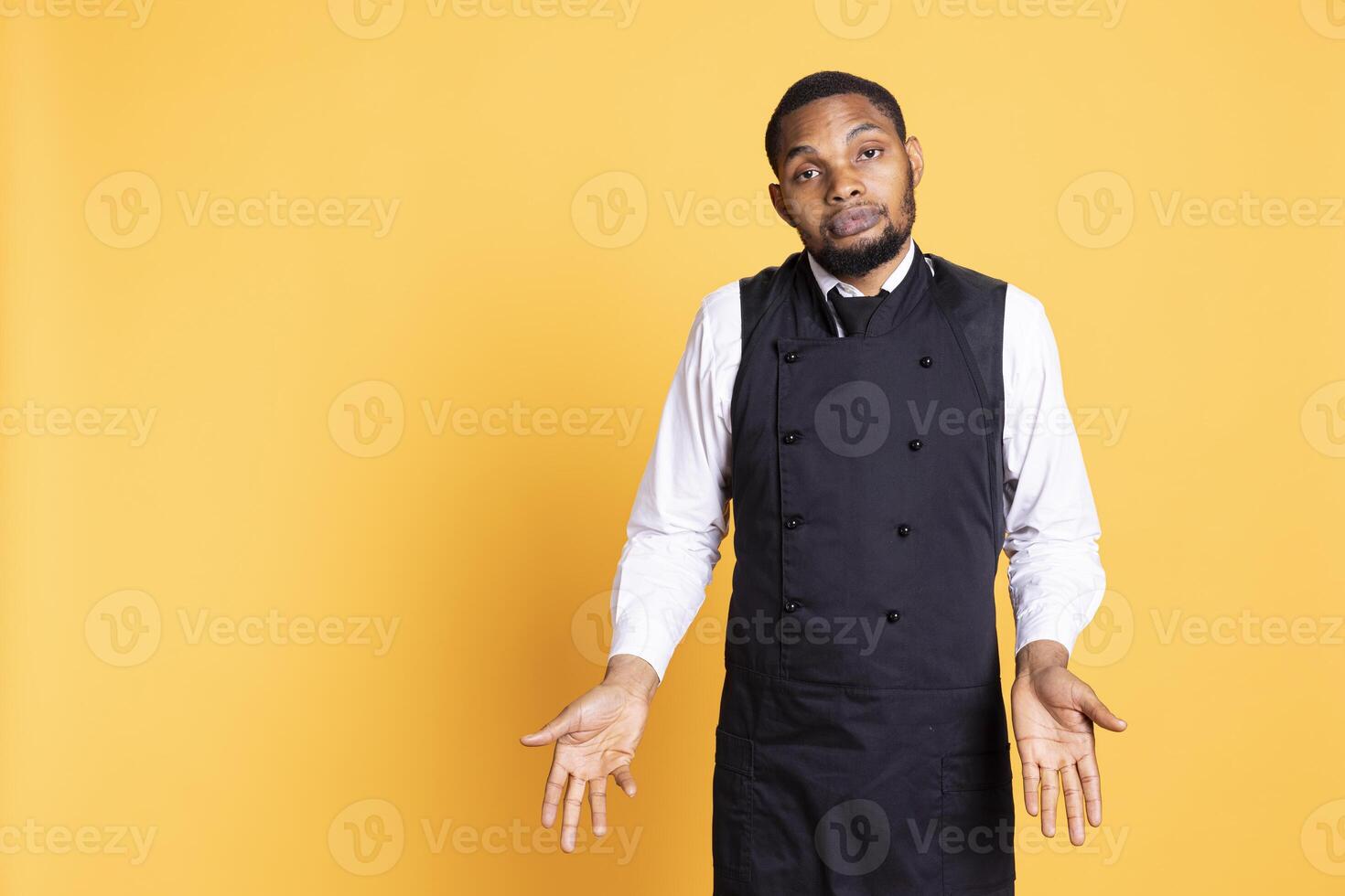 Young butler shrugging and doing i dont know gesture in studio, expressing his uncertainty with a doubtful expression. Restaurant staff waiter in apron acting confused and clueless. photo