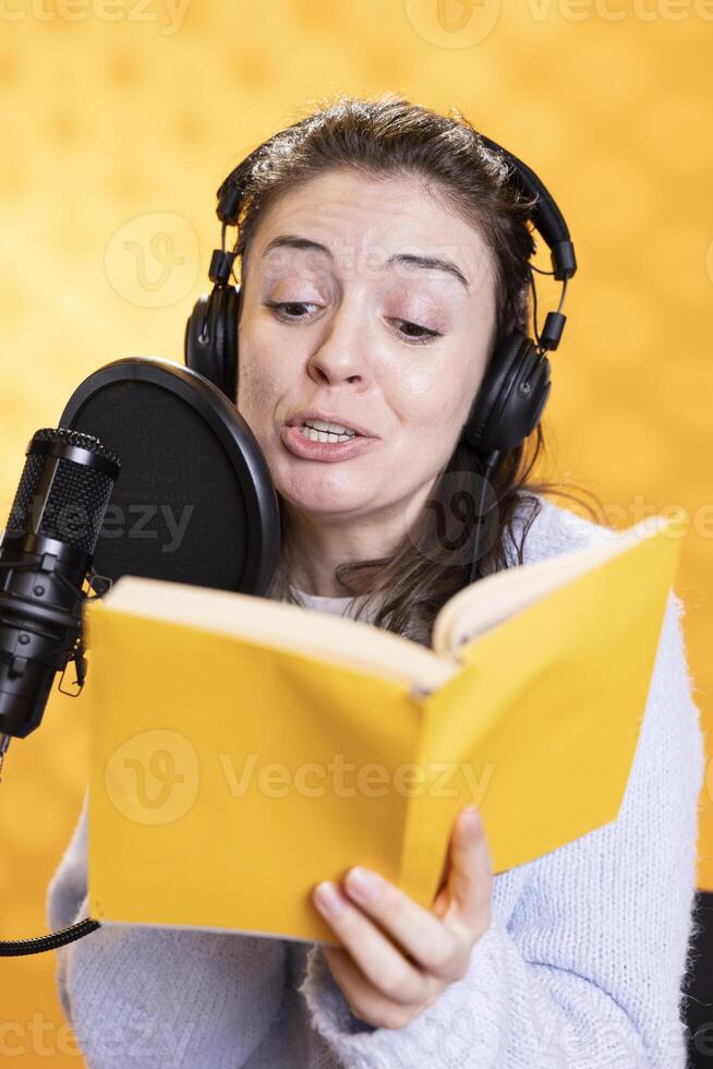 Upbeat voice actor reading kids book, recording audiobook using microphone and headset, studio background. Radiant woman using mic and headphones to produce digital recording of children novel photo