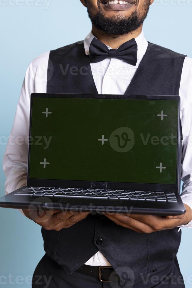 Doorkeeper bellboy holding a laptop with green screen in studio, presenting isolated chroma key display while he works in hospitality industry at a five star hotel. Professional staff. photo