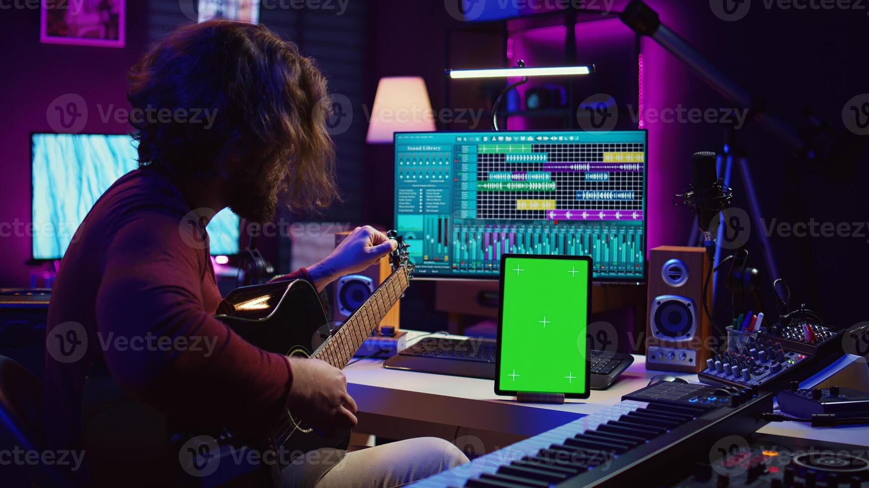 Musical producer learning to tune his guitar before playing, watching lesson tutorial by using greenscreen display on tablet. Young artist tuning strings on instrument, practicing. Camera B. photo