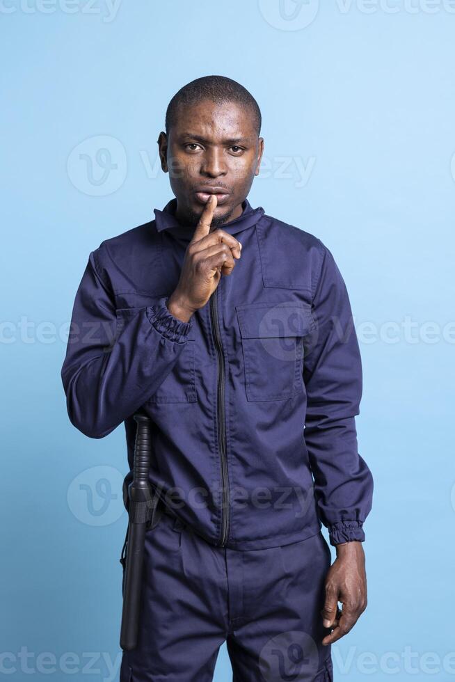 African american patrolling agent puts a finger over lips for silence, doing a hush gesture against blue background. Person in security officer uniform shows mute symbol for a quiet second. photo