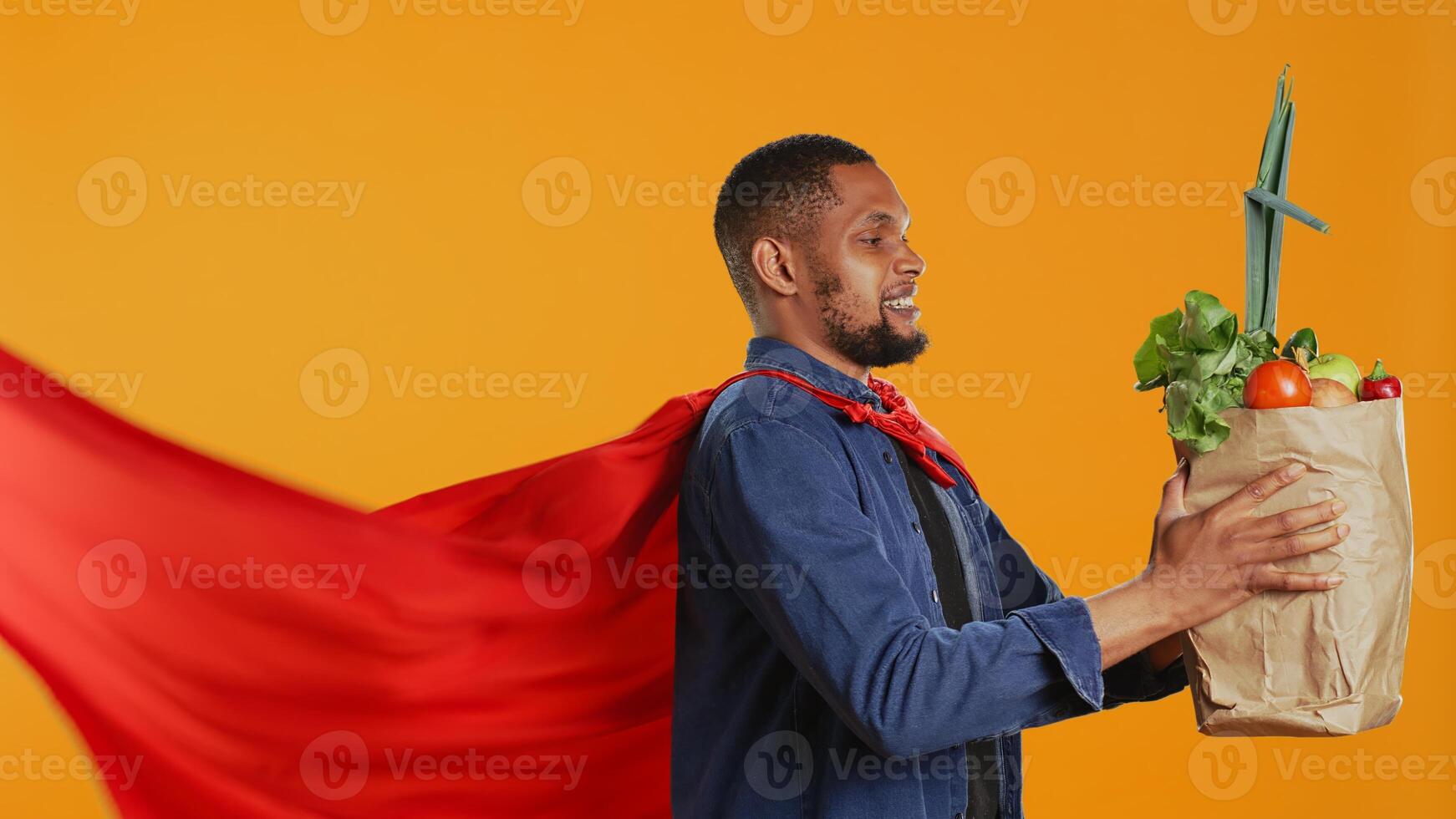 African american guy posing as a superhero with fresh ripe produce, hero with red cape promoting ethically sourced food and zero waste. Male model supports local farming. Camera B. photo
