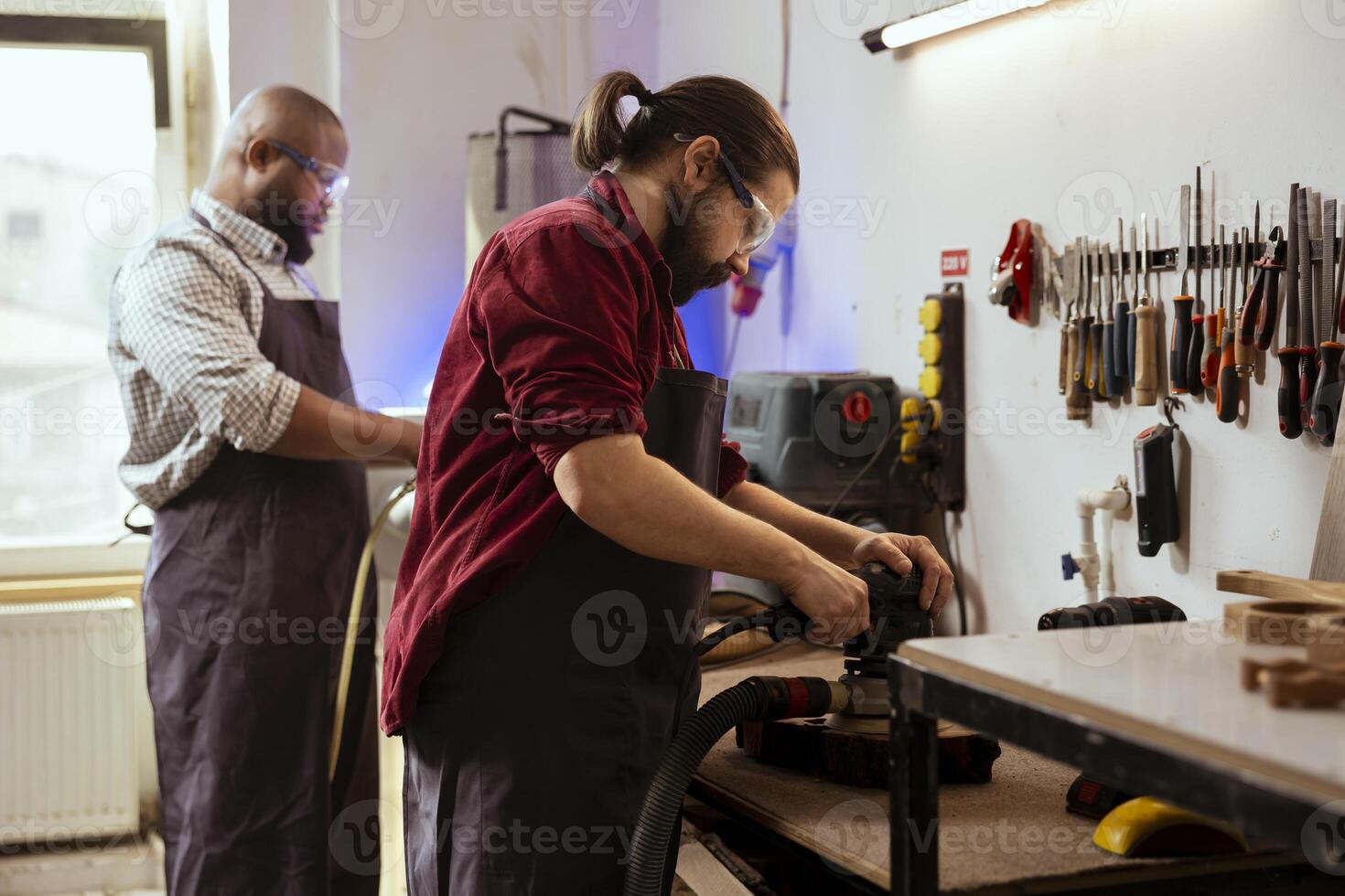Carpenter and BIPOC coworker using angle grinder on lumber block to smooth surfaces by abrasion with sandpaper. Man uses orbital sander gear for furniture assembling, helped by colleague in background photo