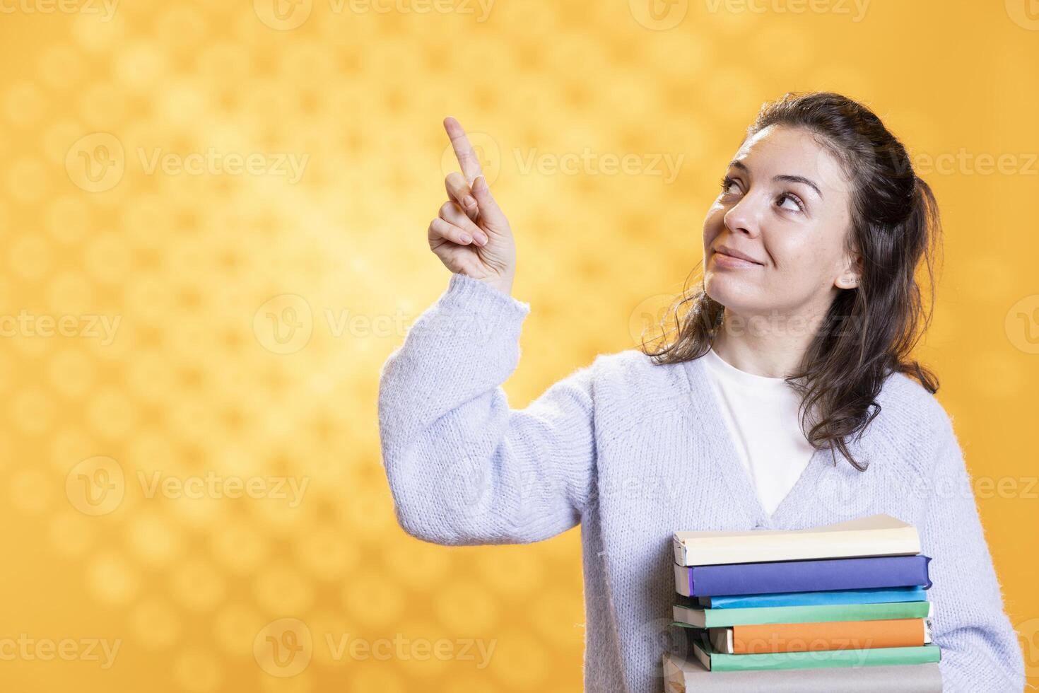 Joyous woman holding pile of books, pointing towards empty copy space, studio background. Cheerful book lover with stack of novels in arms, doing advertisement for literature importance photo