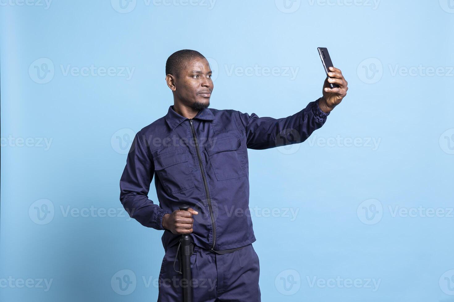 Proud security guard taking pictures with his baton in studio, using a mobile phone to take selfies against blue background. Young bodyguard captures moments in his patrol uniform. photo