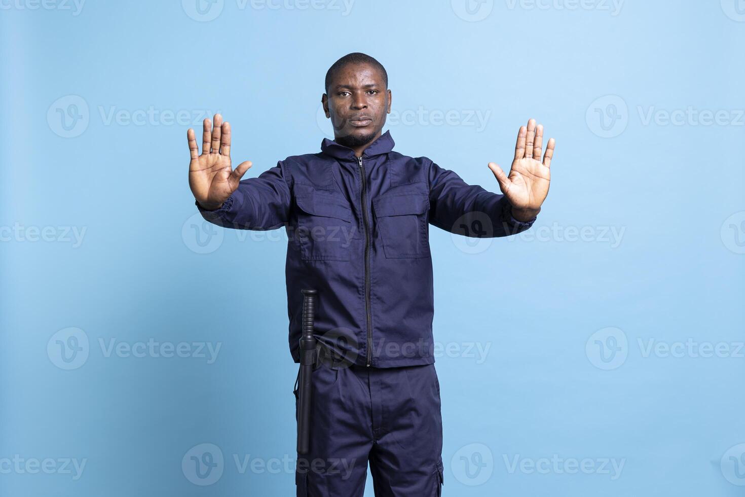 Security guard putting his hands up as a stop sign in studio, creating a forbidden symbol against blue background. Serious agent in uniform restricting the access, does a rejection gesture. photo
