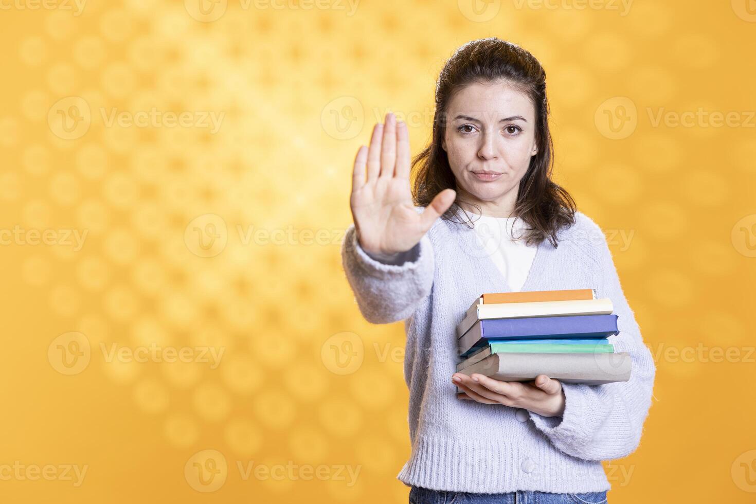 Portrait of woman holding stack of books doing stop sign gesturing, isolated over studio background. Student with pile of textbooks in arms used for academic learning doing halt hand gesture photo