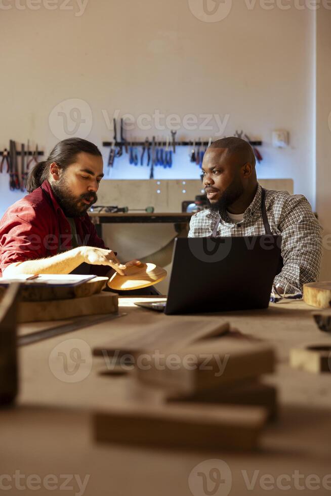 Carpenter and colleague verifying finished wood art product before sending it to client. Artisan and apprentice inspecting wooden object for damages based on laptop checklist photo