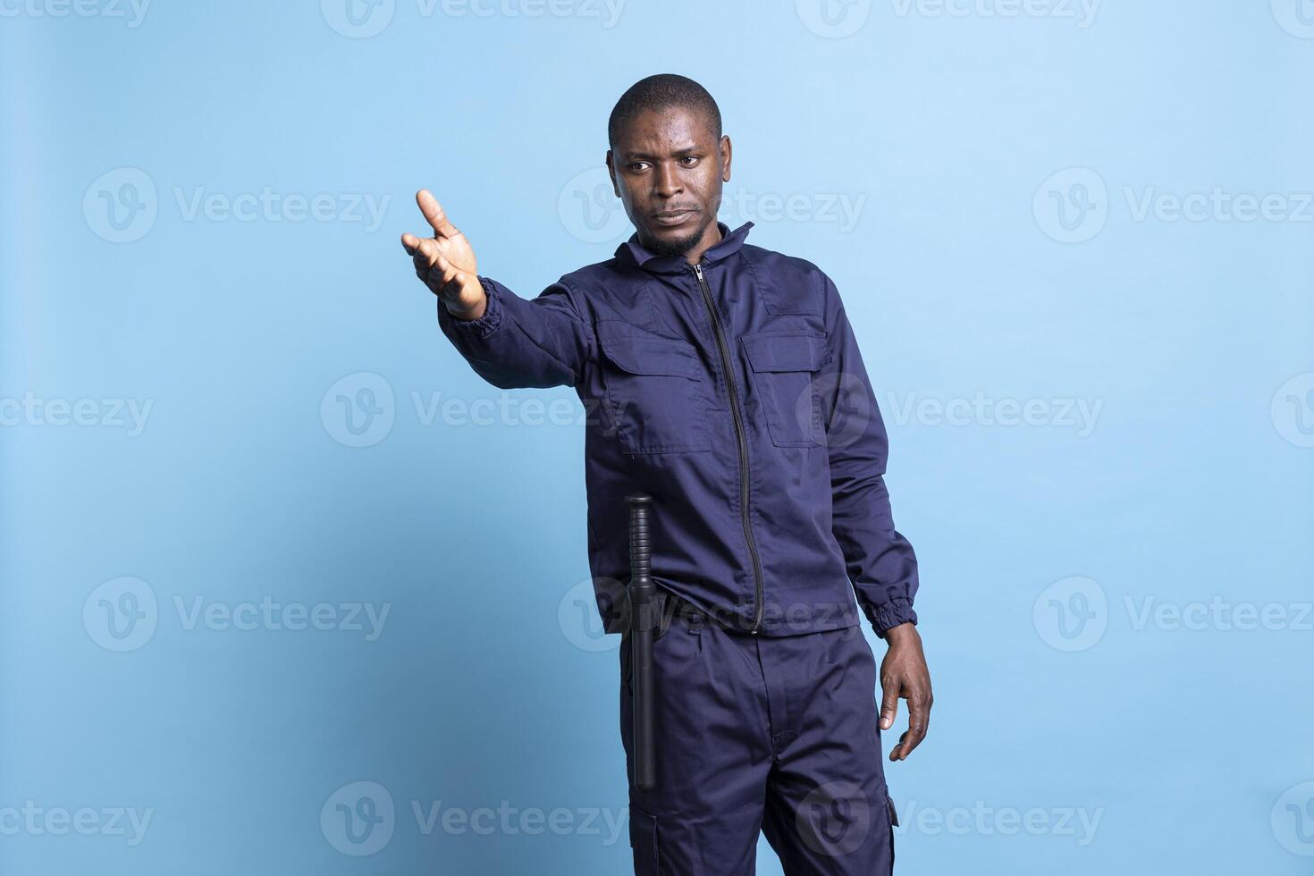 African american security guard indicating the right direction for people, working as a patrol officer against blue background. Bodyguard pointing at an access way to guide the public. photo
