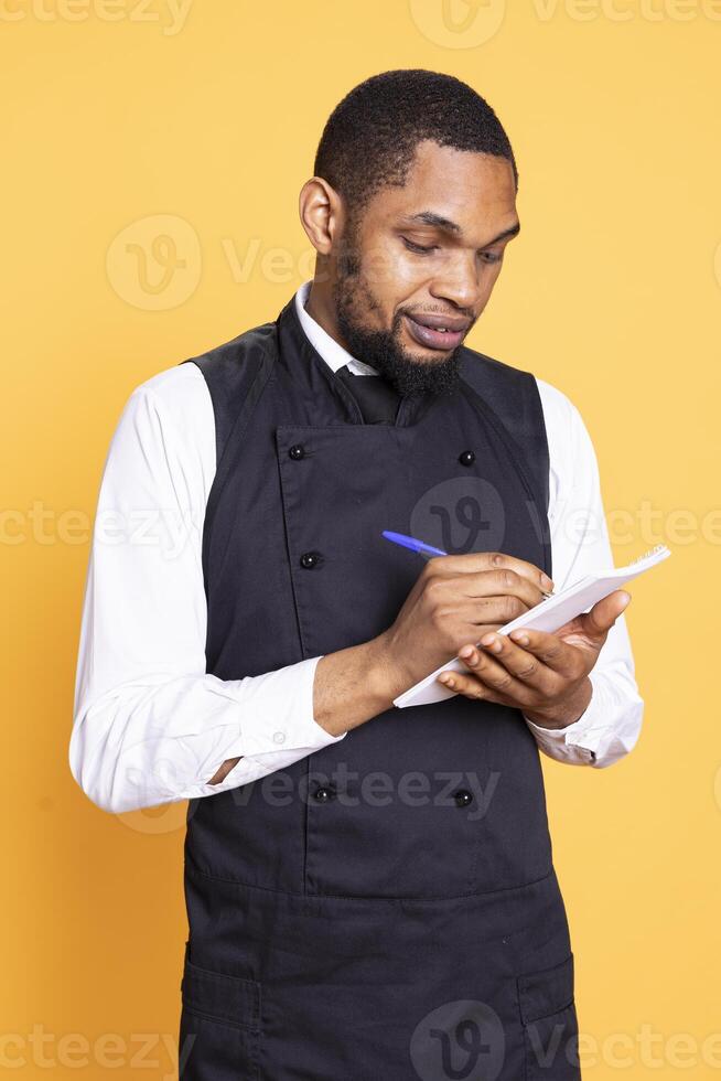 Skilled restaurant server takes the meal order from clients on his notebook, writing the food and drink choice for customer satisfaction. Professional waiter with apron in studio. photo