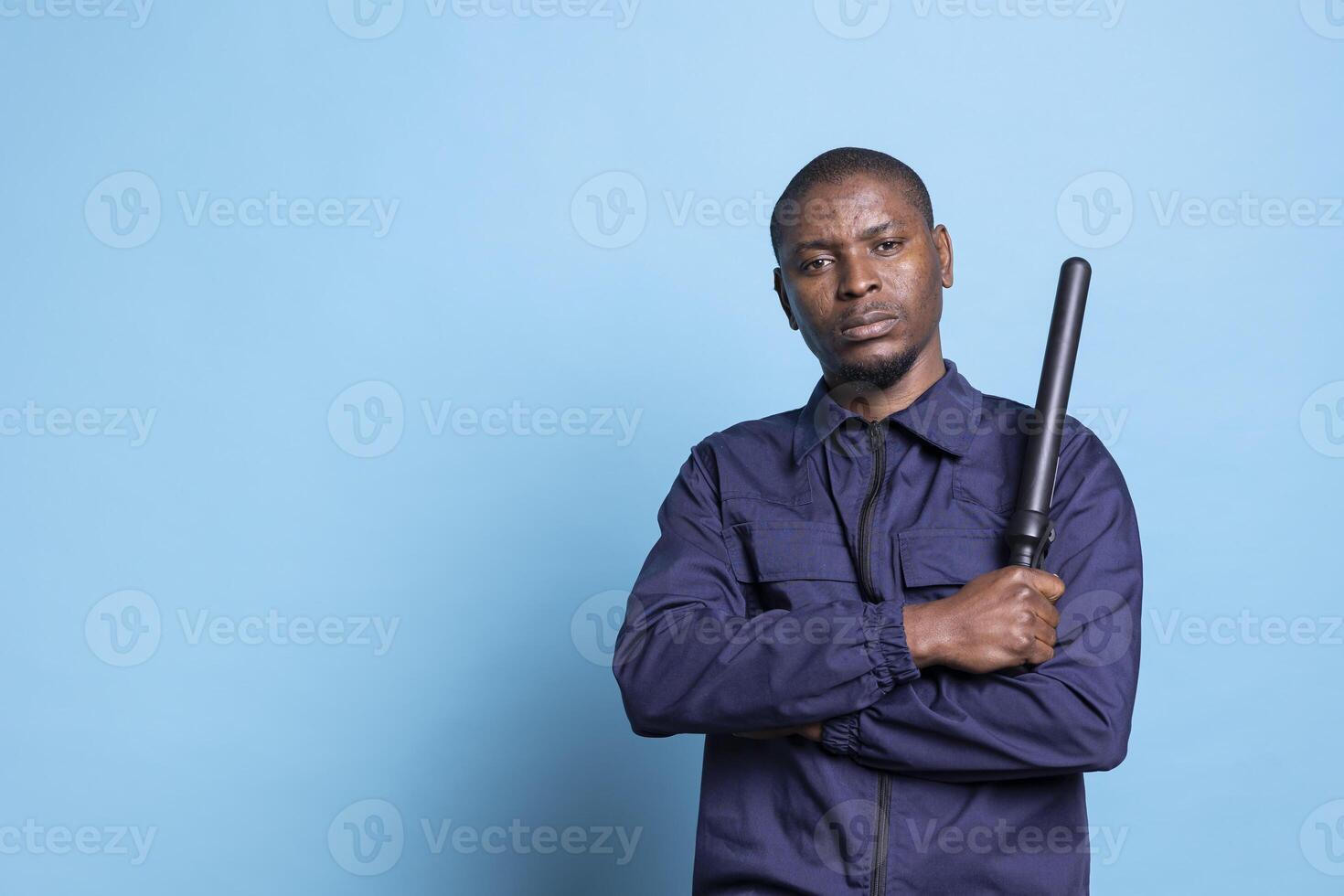 African american security guard feels proud with his baton in studio, posing with confidence with his weapon and professional uniform. Skilled agent standing against blue background. photo