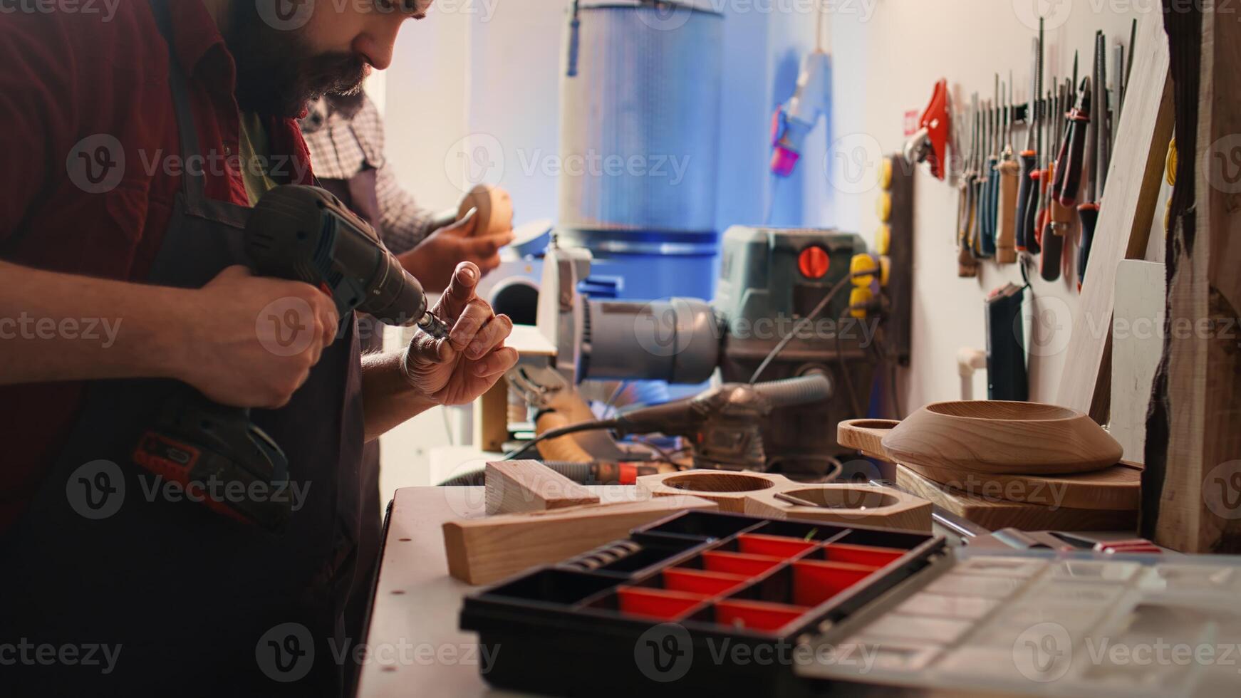 Craftsperson changing power drill head into suitable one for task ahead, making holes into lumber block. Manufacturer in assembly shop picks screw from tool box to use for commissioned work, camera B photo