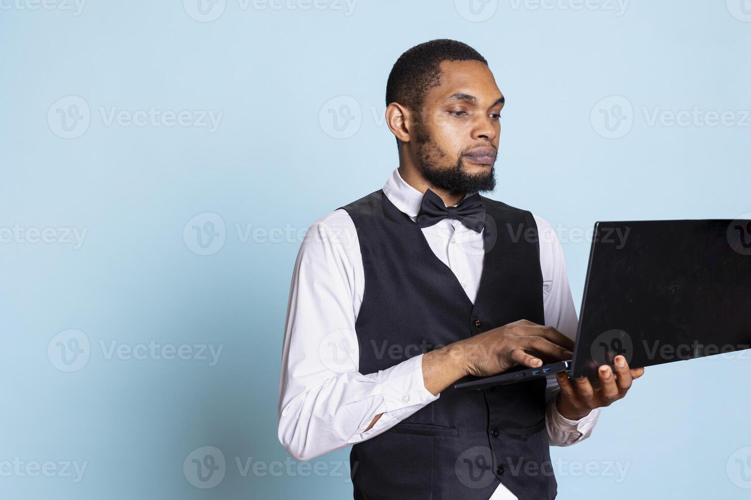 Doorman porter making online room booking reservations on laptop, browsing the hotel website to check lodging availability. Hotel front desk staff gives assistance to guests in studio. photo
