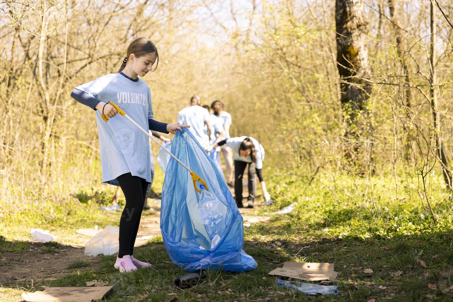 Cheerful proud young kid finishing litter cleanup in a forest, restoring natural environment and helping with forest pollution. Girl recycling plastic waste and using a long claw tool. photo