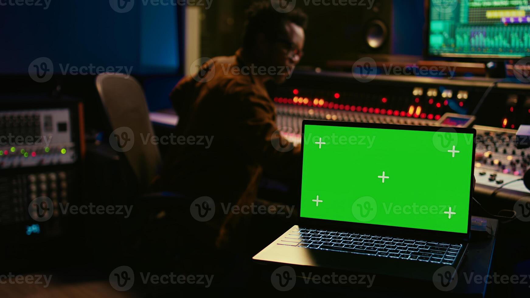 Skilled audio technician processing and mixing sounds next to mockup display on laptop, using variety on knobs, buttons and sliders to edit audio recordings and produce new music. Camera B. photo