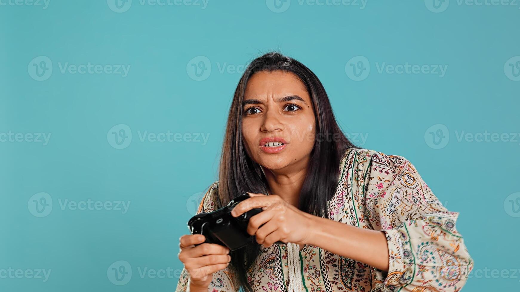 Gamer annoyed after losing online multiplayer videogame, being defeated by rival players. Indian woman upset after seeing game over message, holding controller, studio background, camera B photo