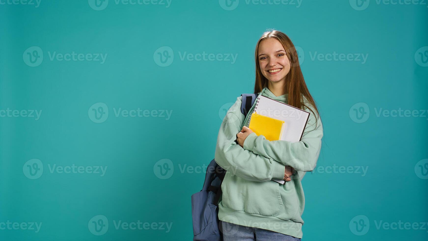 Portrait of joyous teenager with rucksack holding school notebook and notes, isolated over studio background. Jolly pupil with school supplies in arms, preparing to go to college, camera B photo