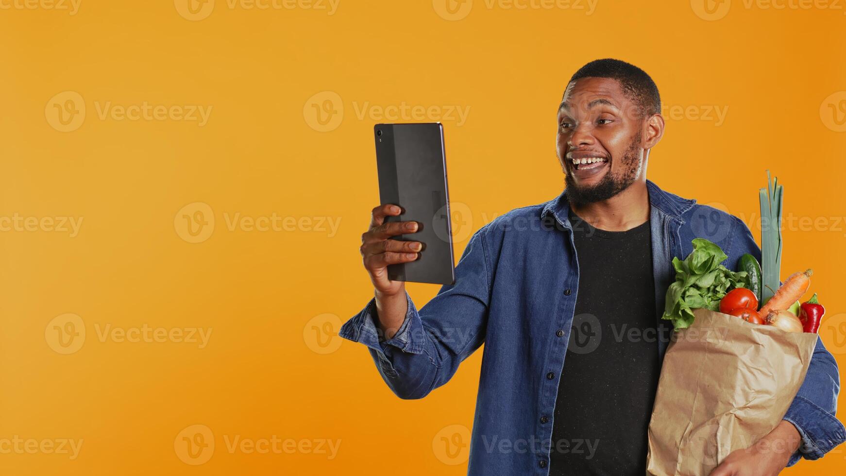 Cheerful guy recommends bio homegrown goods on call, using a tablet to present his groceries from the zero waste eco friendly store. Vegan person talks on teleconference. Camera B. photo