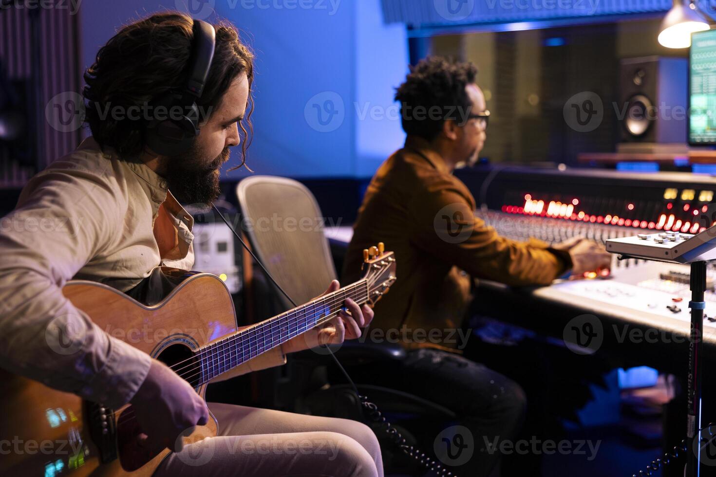 Musician composing new tracks on his electro acoustic guitar, playing record on instrument to create an album. Singer collaborating with producer to make new records in control room at studio. photo