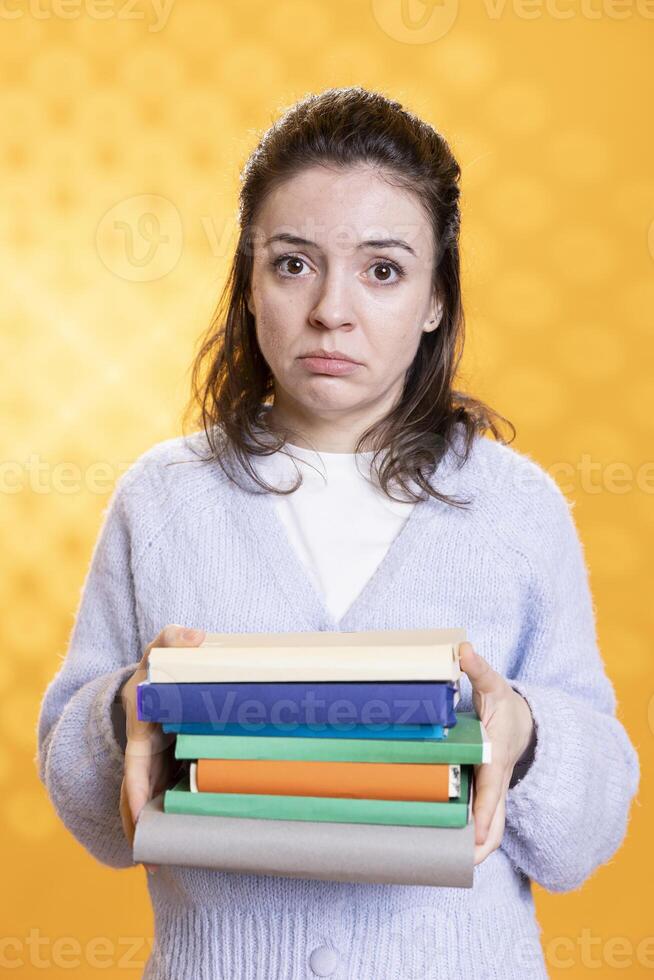 Portrait of woman with distressed facial expression carrying books, afraid of upcoming school exam, studio background. Tense student with textbooks feeling distressed before college assessment photo