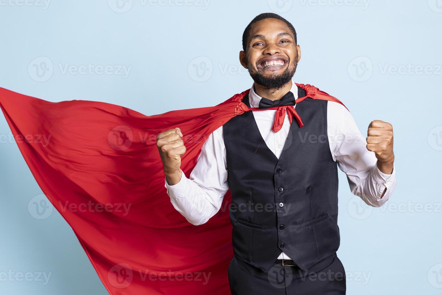 Confident bellboy with suit and tie wearing a red superhero cape, posing as a superhuman and smiling. Young doorkeeper at hotel protecting the guests, hero concept in studio. photo