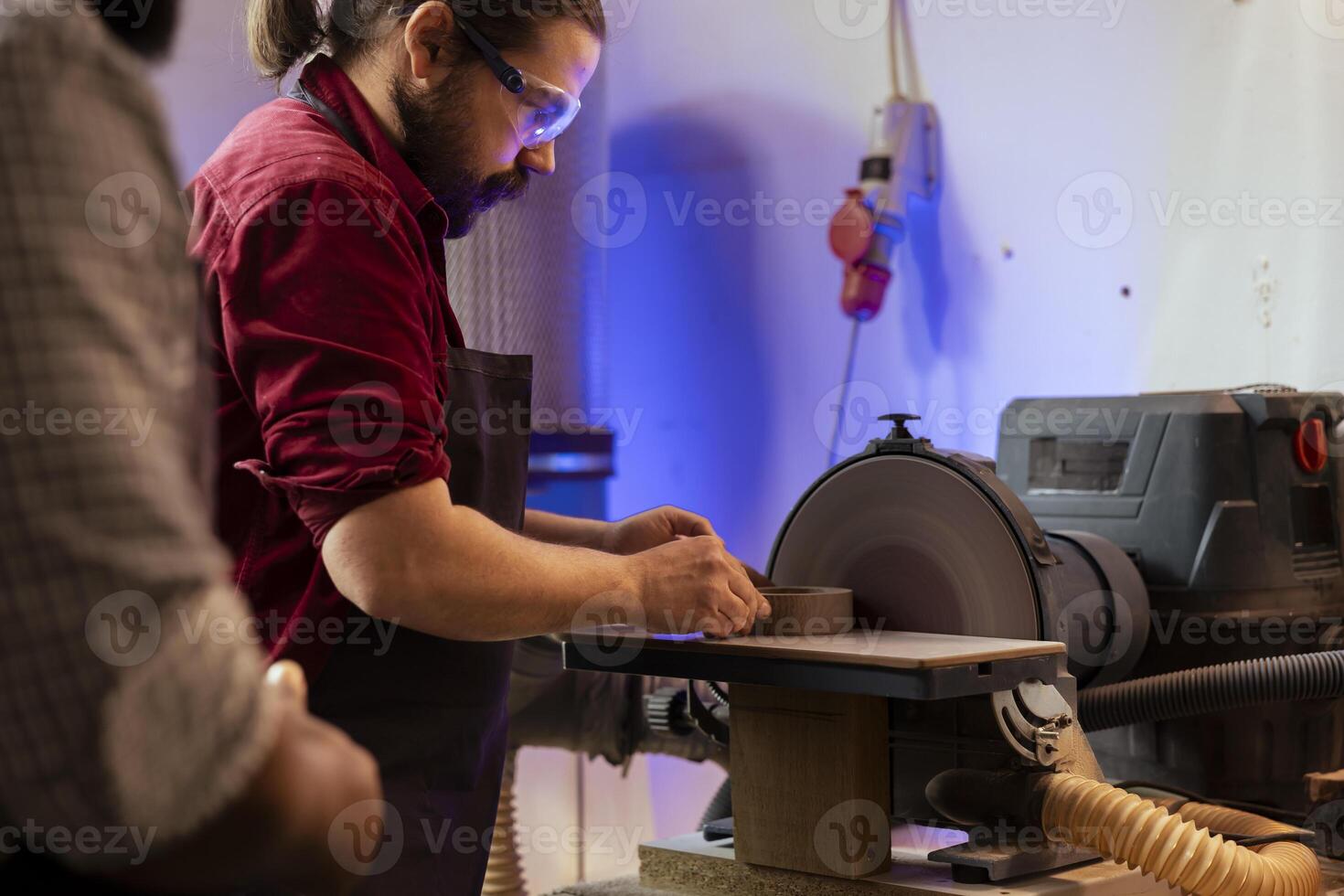 Carpenter in woodworking shop wearing protection glasses while shaping wooden object on disc sander. Cabinetmaker with safety equipment in joinery using professional grinding machine photo
