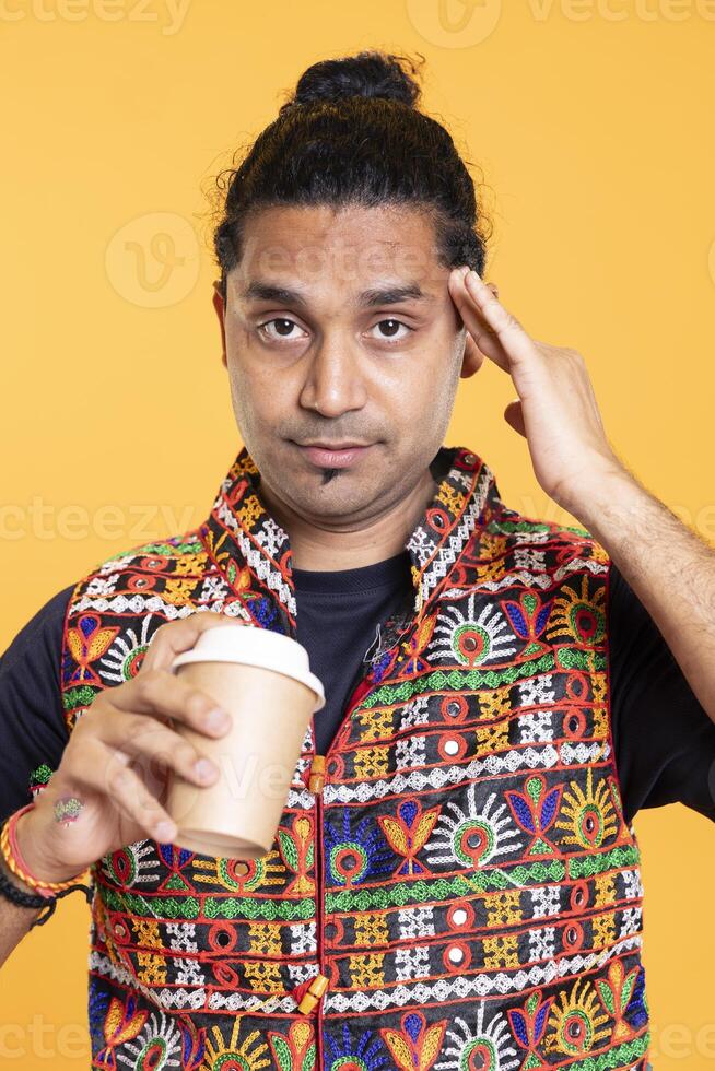 Portrait of sleepy man suffering from headache after exhausting sleepless night, sipping coffee. Hurt indian person rubbing head temples, feeling strain, drinking caffeine beverage, studio backdrop photo