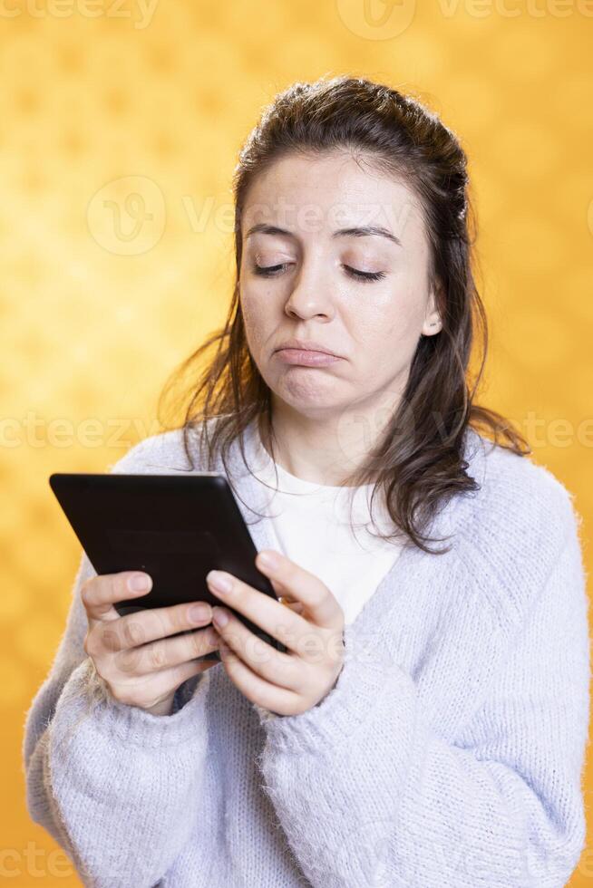 Woman pouting while reading soppy ebook on ereader, enjoying hobby during leisure time. Person with tablet in hands glooming, upset by character death in digital novel, studio background photo