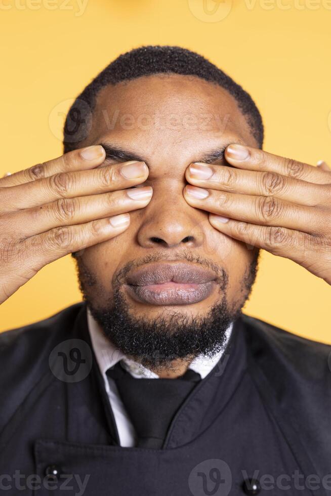 Restaurant worker showing the three wise monkeys gesture in studio, covering his eyes and see no evil as a sign of wisdom and concealment. Butler in apron shows wise symbolic metaphor. photo