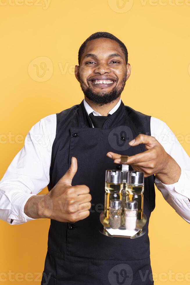 Catering employee showing a thumbs up gesture in studio, presenting his approval and content emotions on camera. Young restaurant waiter gives a like sign, professionalism. photo