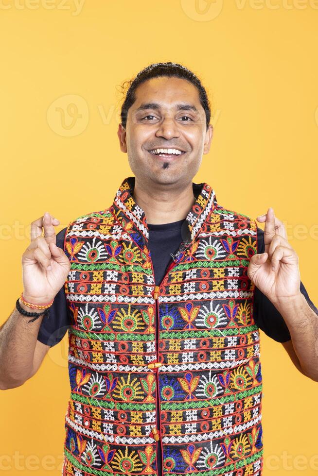 Portrait of joyful hopeful man crossing fingers, wishing to receive good news, studio backdrop. Upbeat person with positive facial expression hoping for good luck, aspiring for miracle photo