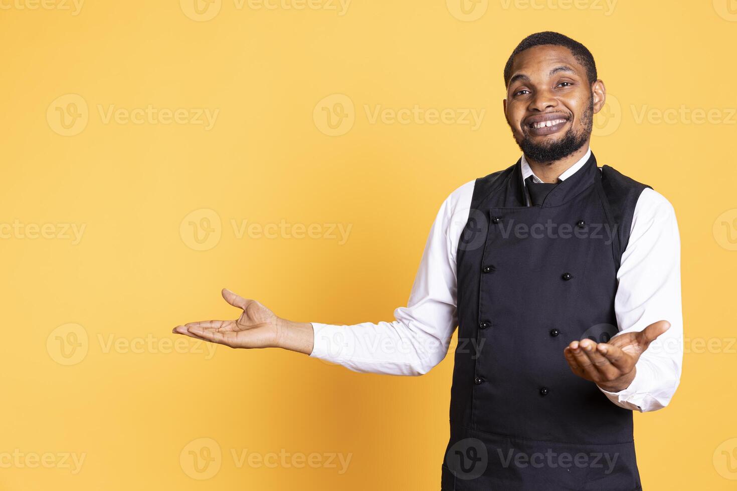 African american waiter points at something to his left or right, indicating direction to the sides in studio. Customer focused employee wears uniform and apron against background. photo